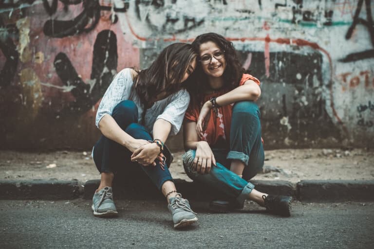 Två kvinnor som är bästa vänner och som sitter på en trottoar framför en vägg med graffiti