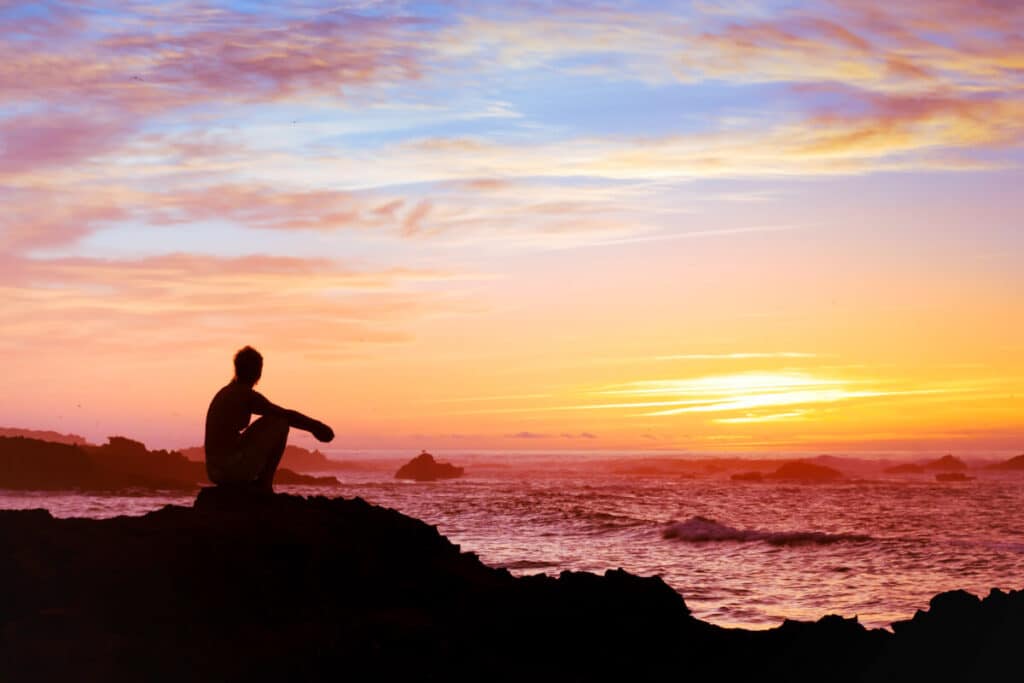 Kvinna som sitter ensam vid solnedgången nära havet och funderar på vad som är meningen med livet
