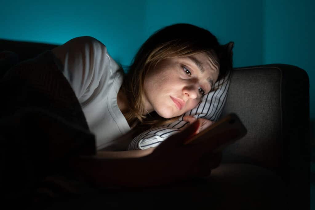 Ung kvinna som ligger i soffan och surfar i mobilen samt undrar hur man stänger av hjärnan när man ska sova